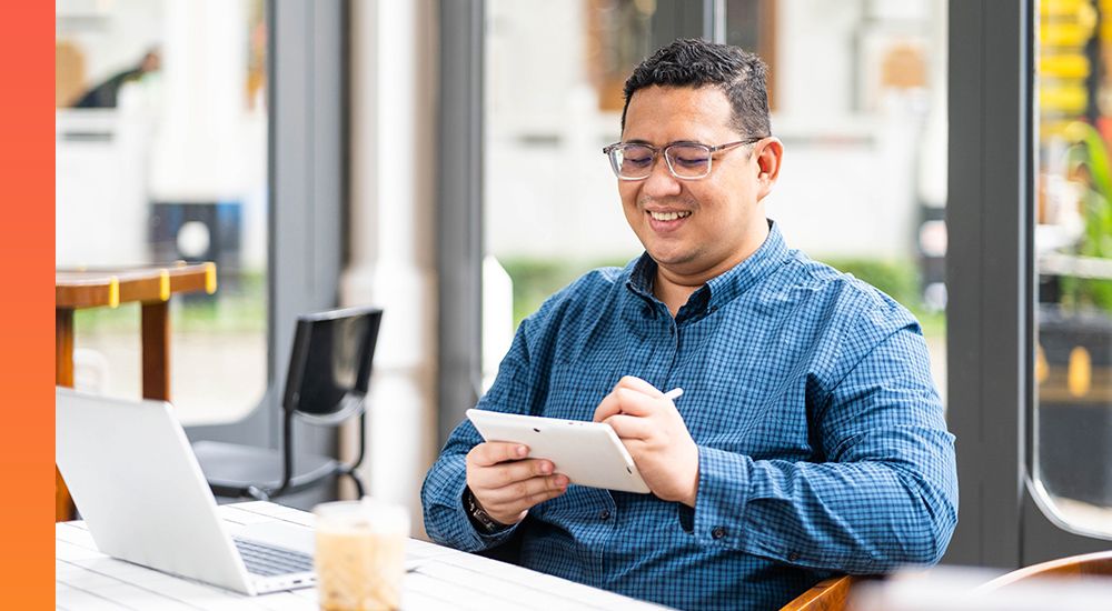 Man in a blue shirt sitting at a desk with a laptop in front of him while working on a tablet device 