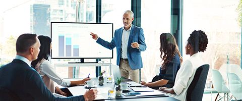  Man in a blue suit jacket standing in front of a screen with graphs while presenting to people at a conference table