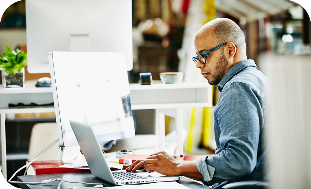 Man in a gray shirt sitting at a desk while working on the laptop in front of him