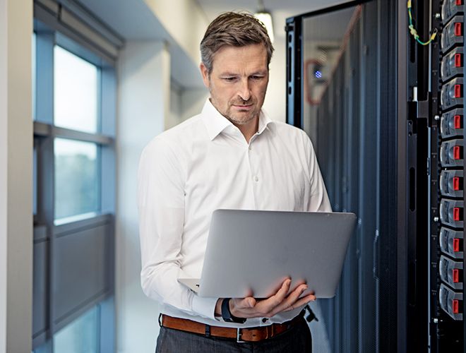 Man standing next to a server stack while looking at a laptop he's holding