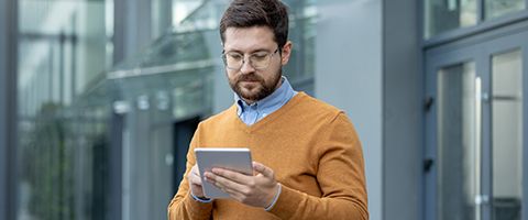 A person in business casual stands outside a building and uses a tablet.
