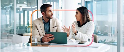 Two colleagues sit at a table in a modern office, discussing information on a tablet inside a digital orange box with rounded corners.