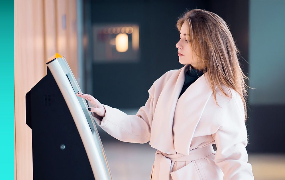 A person with long hair wearing a light-colored coat standing in front of and interacting with hospitality software on an electronic kiosk.