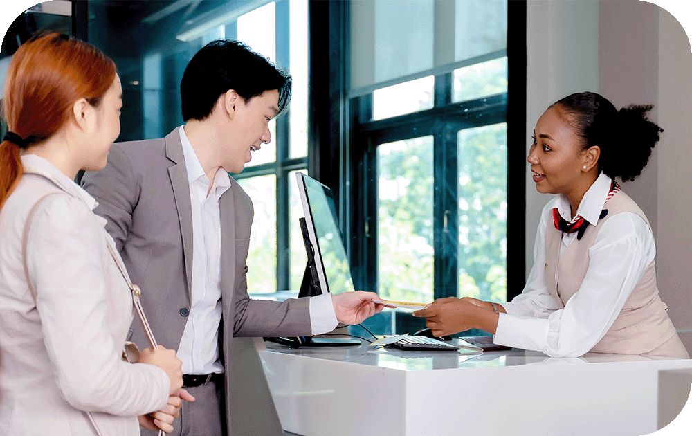Two individuals are standing at a hotel reception desk  while a person behind the desk at a computer monitor hands them a document. 