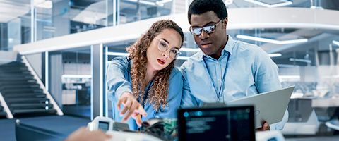 Two professionals in glasses and light blue button-ups collaborate on a project and look at a computer together