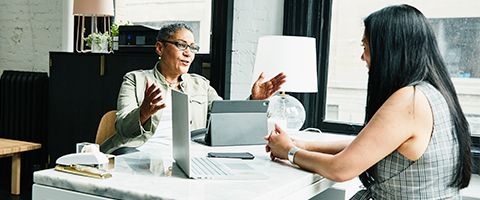 Two people in business casual attire talking and sitting across from each other at a desk with a laptop and smart tablet.