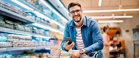 A person in glasses and a jean button-up at a grocery store stands in a food aisle with a cart and phone in their hands while looking up and smiling