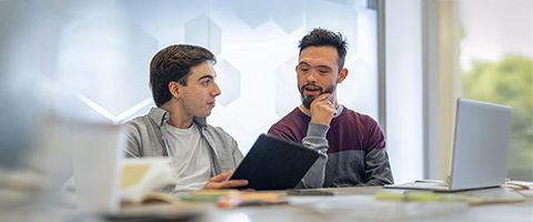 Two coworkers sit next to each other at a table looking at the same tablet and discussing work