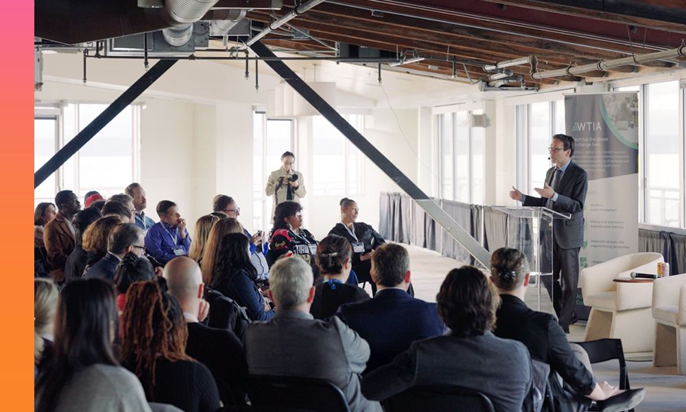 A group of people seated in a room with large windows listens attentively to a speaker at a podium next to a banner displaying the acronym WTIA. 