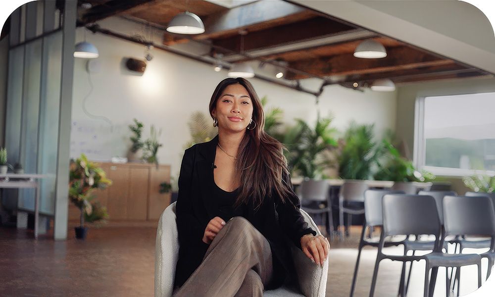 AI2 Incubator Events & Community Manager Audrey Yun sits on a chair facing forward in a modern, well-lit room with wooden ceilings, large windows, potted plants and hanging lights.