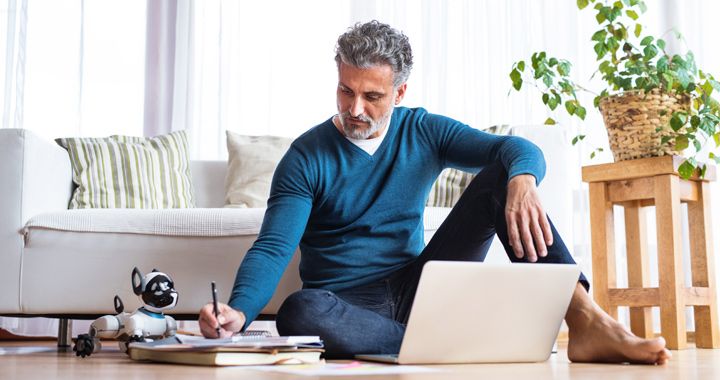 Hombre sentado en el piso de su hogar trabajando en una computadora portátil mientras toma notas