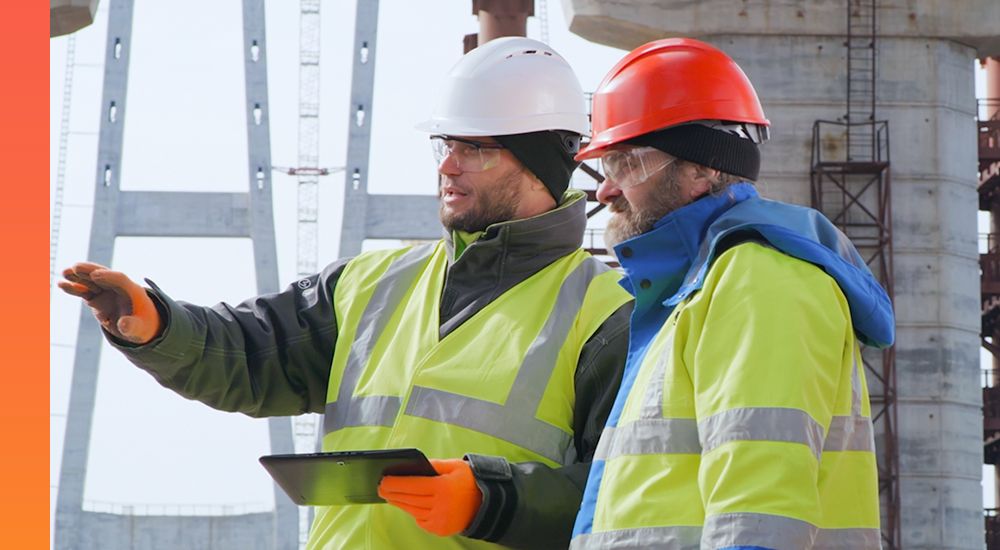 Two men in yellow safety vests and hardhats looking at computer tablet and point out solutions at construction site. 