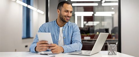 A professional sits at their desk with a tablet in their hands while looking and smiling at their laptop