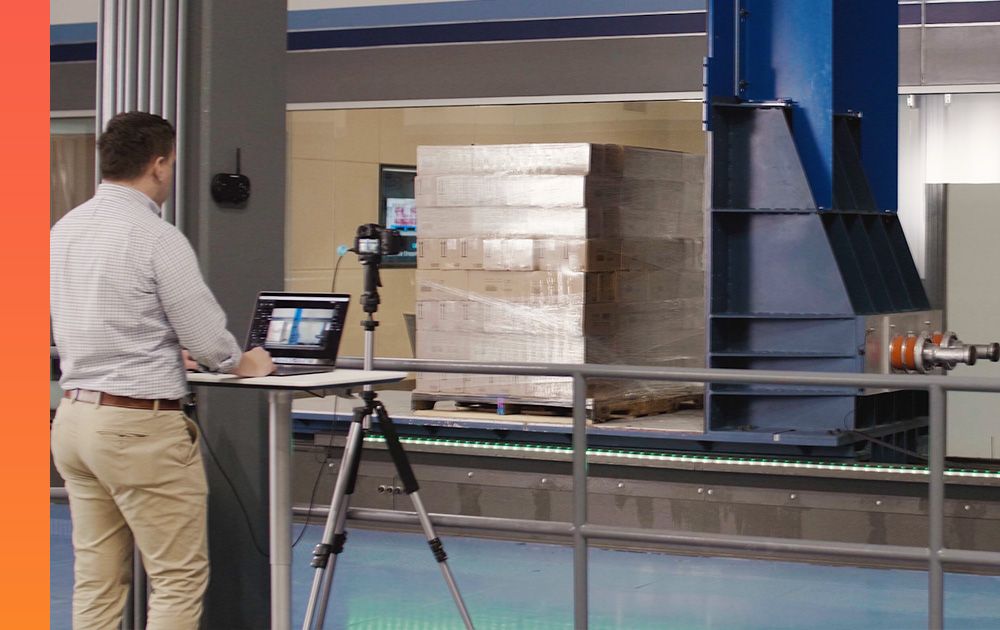 An individual works on a laptop while standing in front of a pallet of goods on an industrial platform.