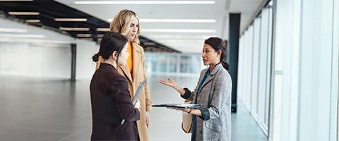Three people in business attire talk in front of a long wall of windows.