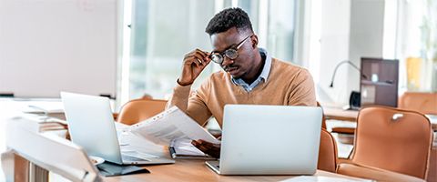 A person reads a document at a table and touches the rim of their glasses in a bright room.