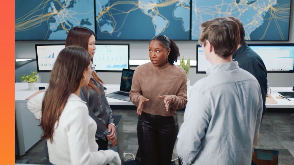 A group of five people talk in a room with computers on tables and a map on the wall.