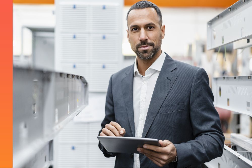 A person in a business suit is holding a tablet while standing in a warehouse, surrounded by shelves and inventory