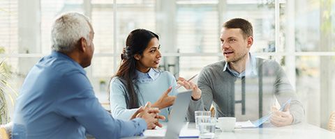 Three people sit at a table in a bright office and talk.