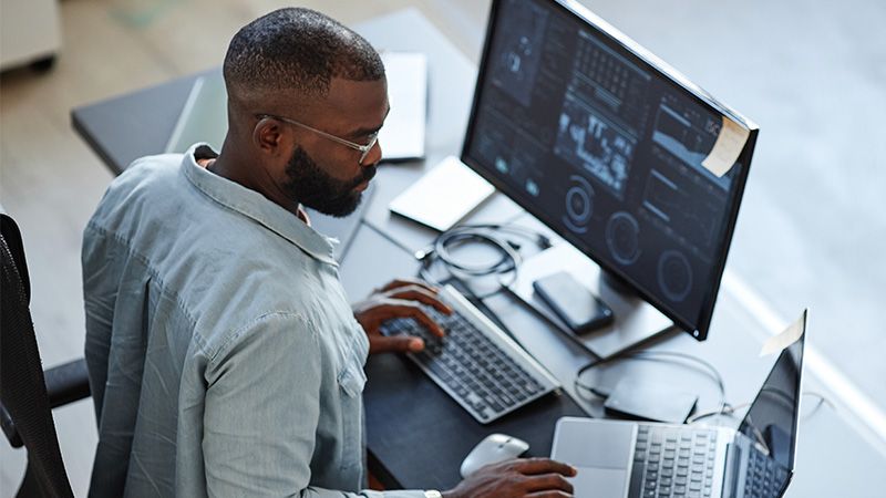 Draufsicht eines Mannes, der in einem Büro an einem Laptop und einem Desktop-Computer arbeitet