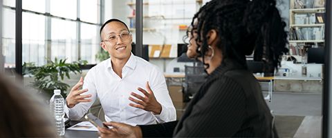 Person in light business attire sitting in an office setting talking to another person in dark attire.