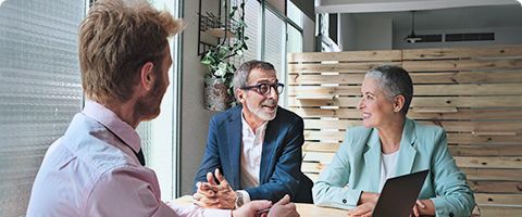 Three people in business attire sit at a table near a bright window and talk.