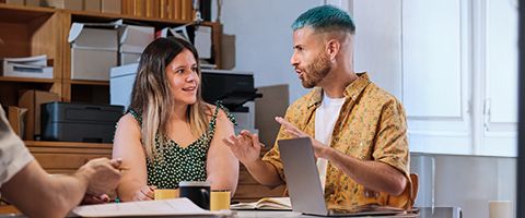 Two people in casual attire sitting at a table and talking next to a laptop.