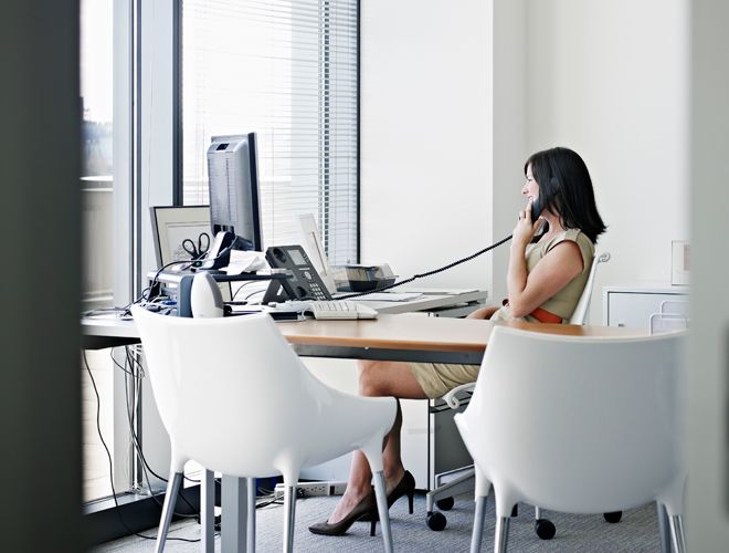 Geschäftsfrau sitzt an einem Schreibtisch und telefoniert vor einem Computermonitor neben einem Fenster