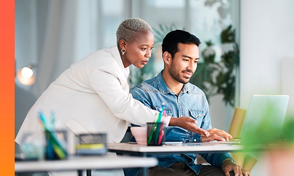 Two individuals at a desk focused on a laptop screen, discussing or explaining something