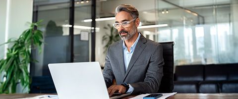 Business professional wearing a suit and glasses works on a laptop in a modern office.