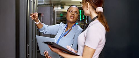 Two IT professionals in a server room examine a laptop, with one person pointing to something offscreen while looking at their colleague.