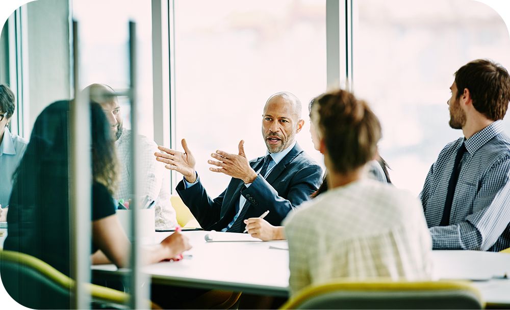 A half dozen people meeting in a conference room while a man in a suit is talking