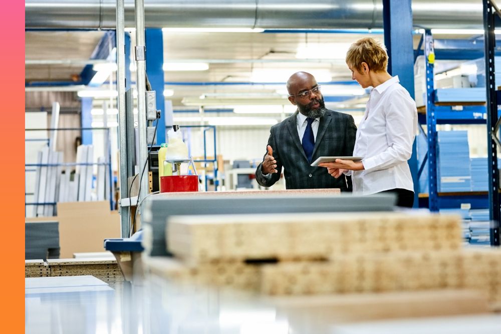 Business man in a suit talking to another person holding a tablet device on a factory floor