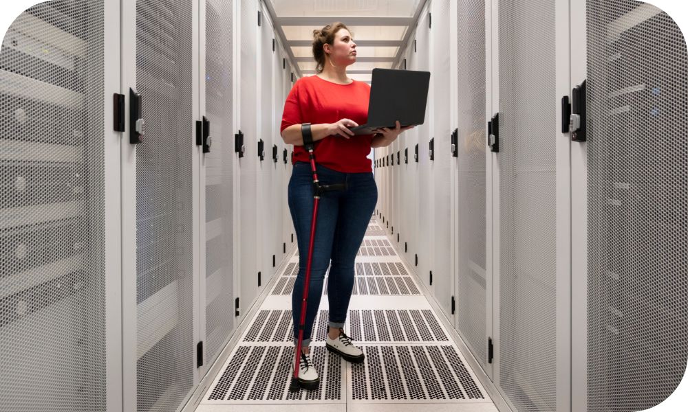 Woman in a red shirt and blue jeans standing in a row of servers while holding a laptop device