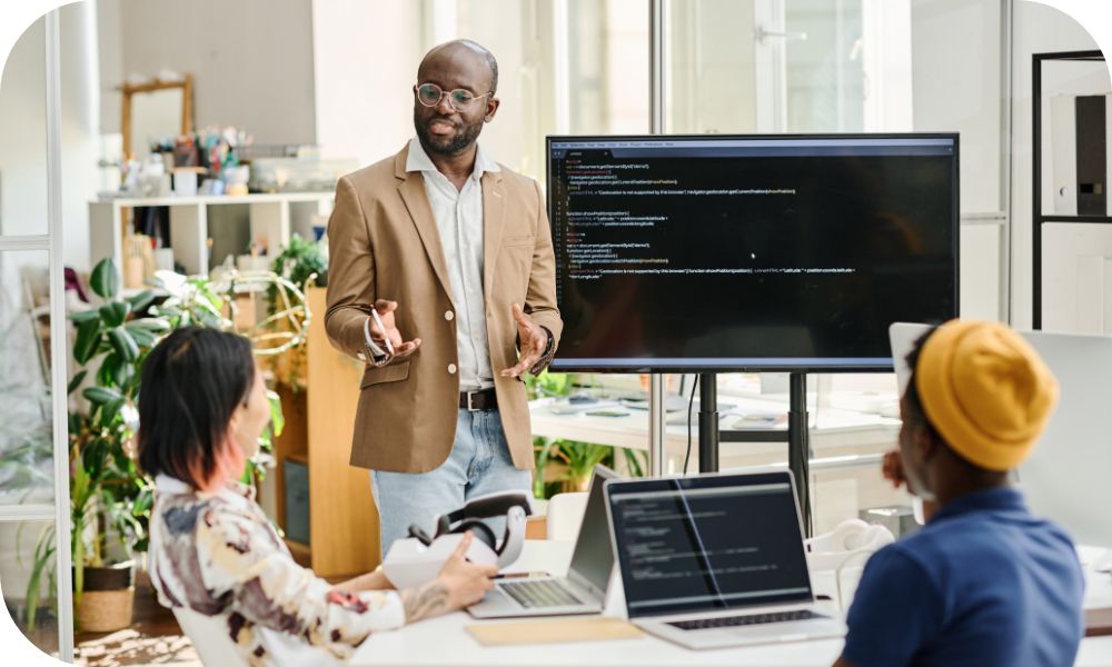  Man standing in front of a screen while talking to two other people sitting down with laptop devices