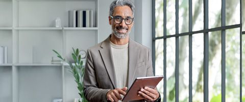 A person in a suit holds a tablet and smiles into the camera.