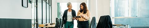 Two professionals stand in an office environment talking and looking at a tablet together.