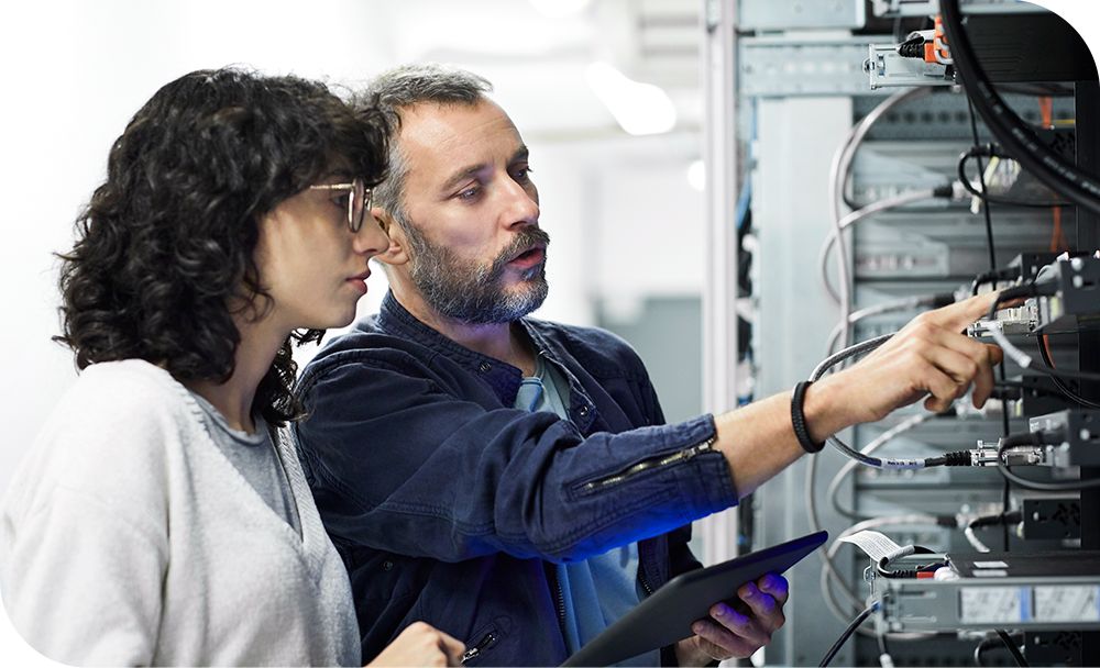 Woman standing next to a man holding a tablet device while pointing towards the server stack in front of them