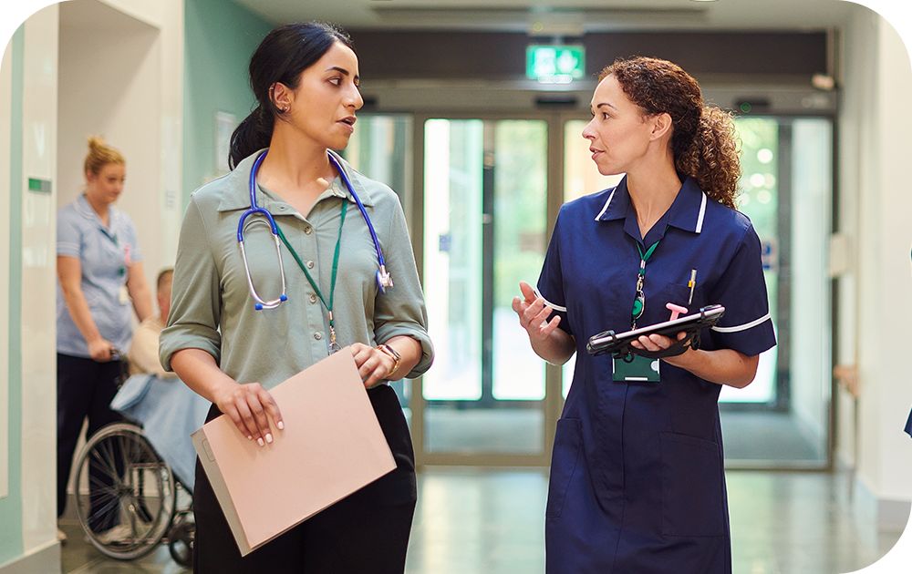 Two healthcare professionals with stethoscopes and medical chart walking and discussing in hospital hallway.