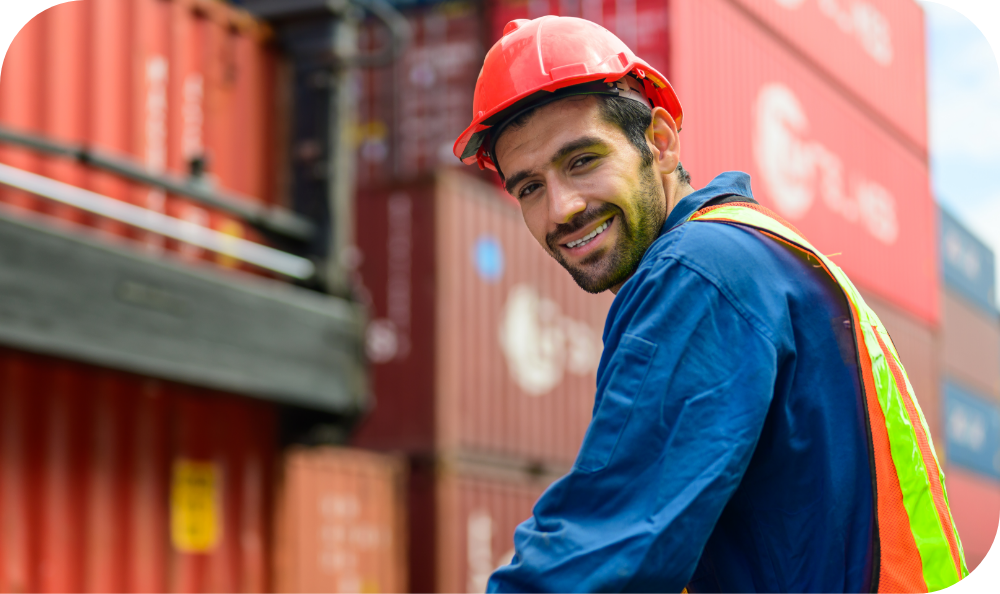 Man in hardhat standing in front of orange shipping containers looking directly at camera over his shoulder.