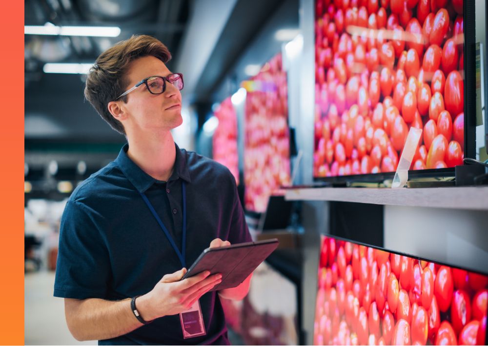 : A professional in a dark blue polo looking up at a television screen on a shelf and taking inventory on their iPad. 
