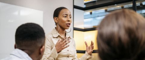 A person gestures while speaking to two individuals in a room with a large screen displaying content