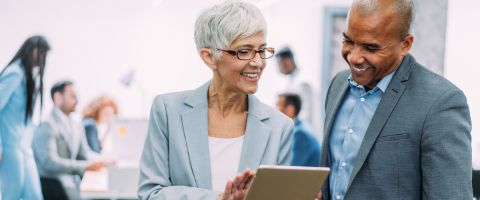 A person in a business suit holds a tablet and another person looks at it while a group of people in the background has a discussion around a whiteboard 