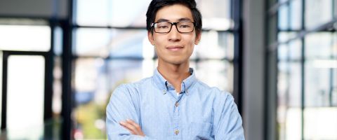 A person wearing a light blue shirt stands in front of a blurred background featuring large windows