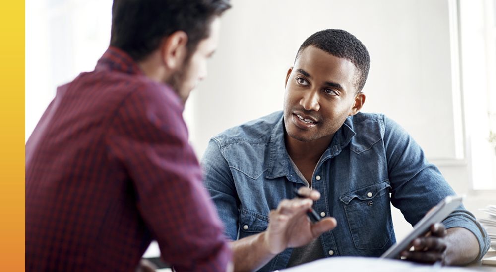 Two individuals engaged in a discussion across a table, one holding a tablet and gesturing with their hand to the other person who is looking at the device