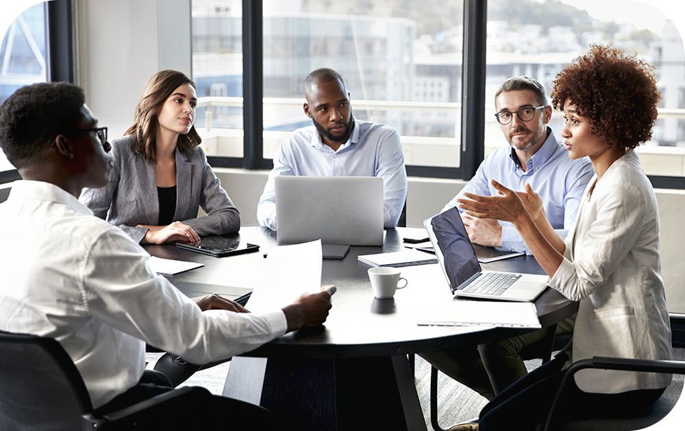 Group of business colleagues focusing on person at head of table explaining issues while others look on intently.