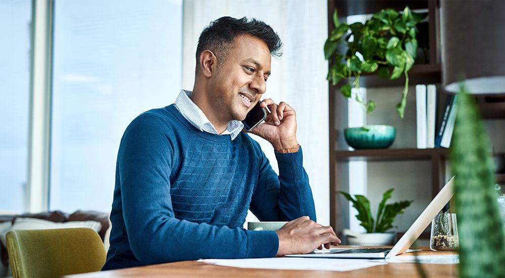 Man in a blue sweater sitting at a desk while talking on the phone and working on a laptop device