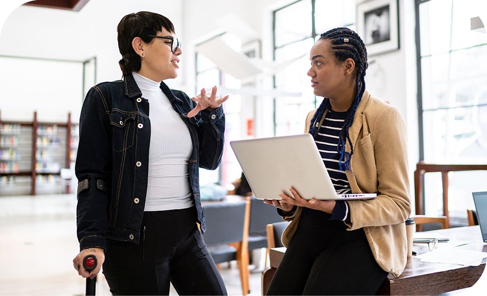 Woman standing with a cane while talking to another woman sitting with a laptop device