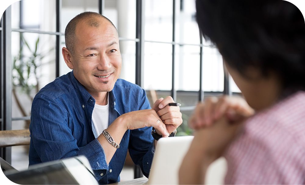 Man sitting at a table in front of a laptop device while talking to someone sitting across from him