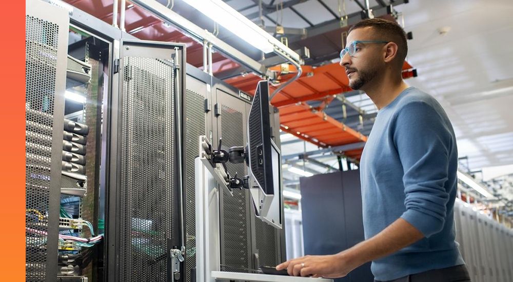 Person in a blue shirt and glasses standing at a computer in a data center. 